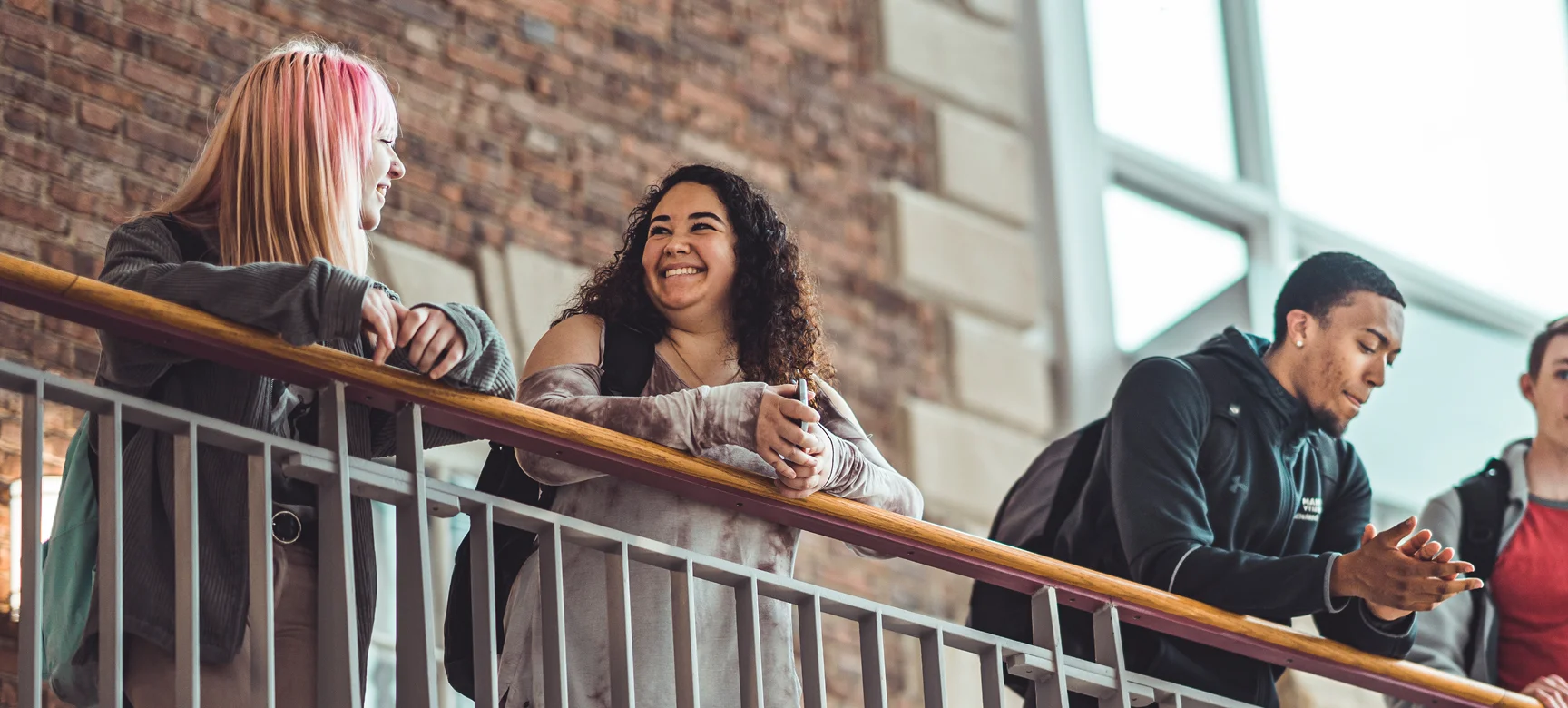 Group of students standing along a balcony railing indoors, smiling and chatting between classes