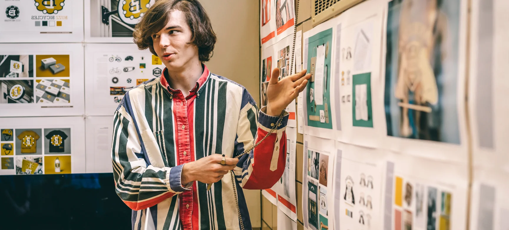Student presenting design work pinned on a wall, gesturing toward visual materials