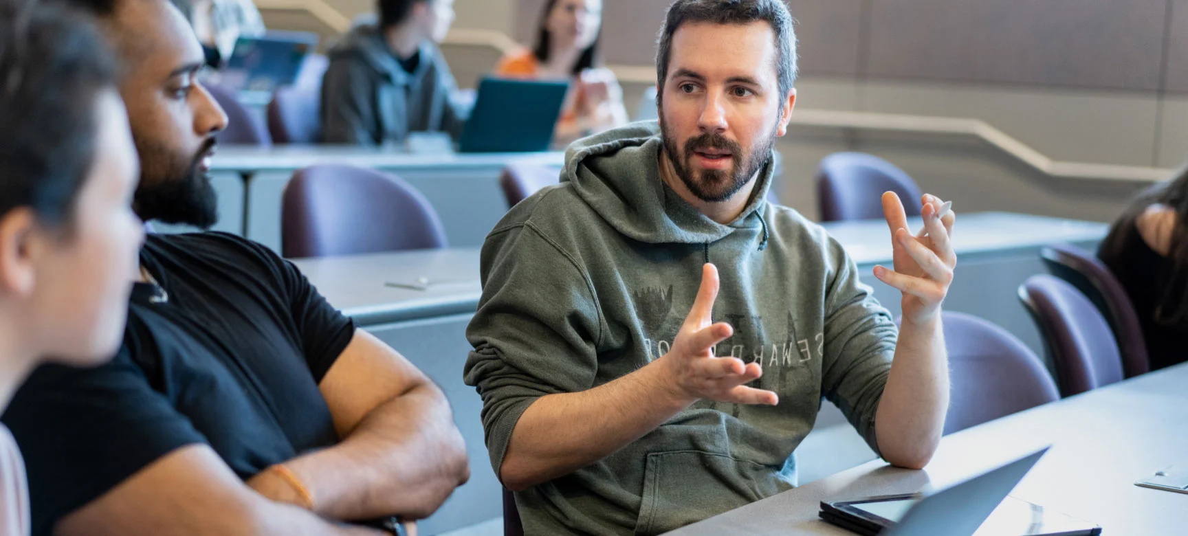 Student in a hoodie speaking and gesturing while talking with classmates in a lecture hall