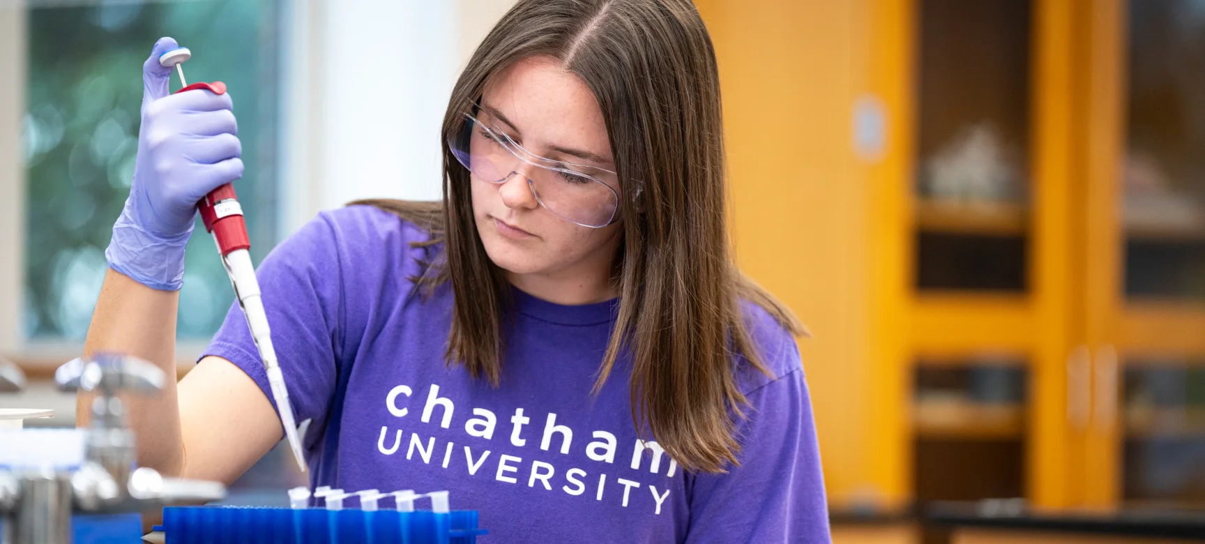 Student in safety glasses and gloves using a pipette in a lab, wearing a Chatham University shirt