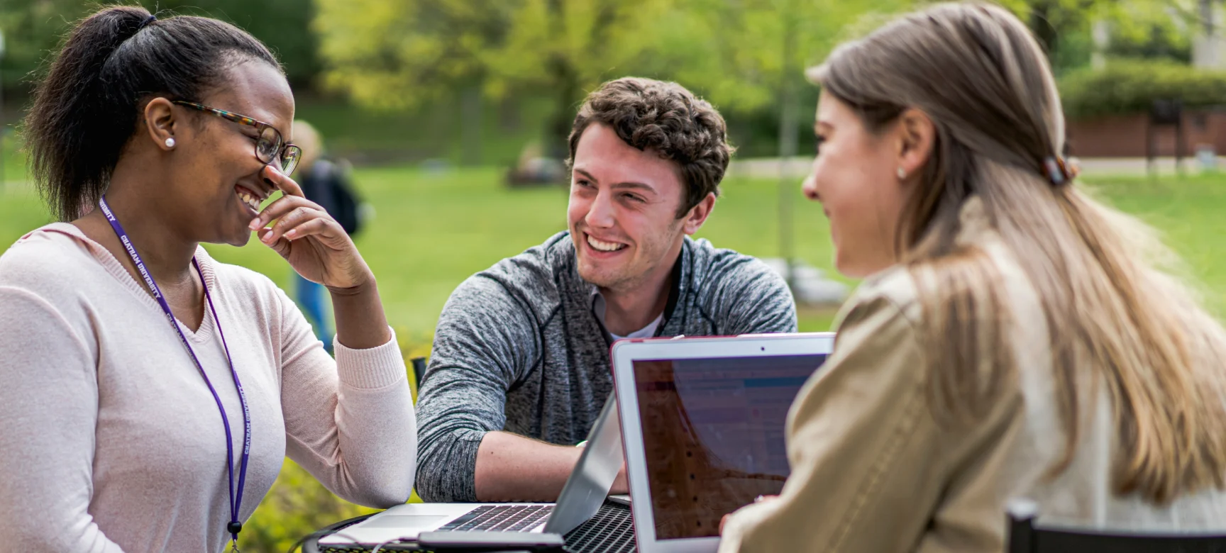 Three students sitting outside with laptops, laughing and chatting