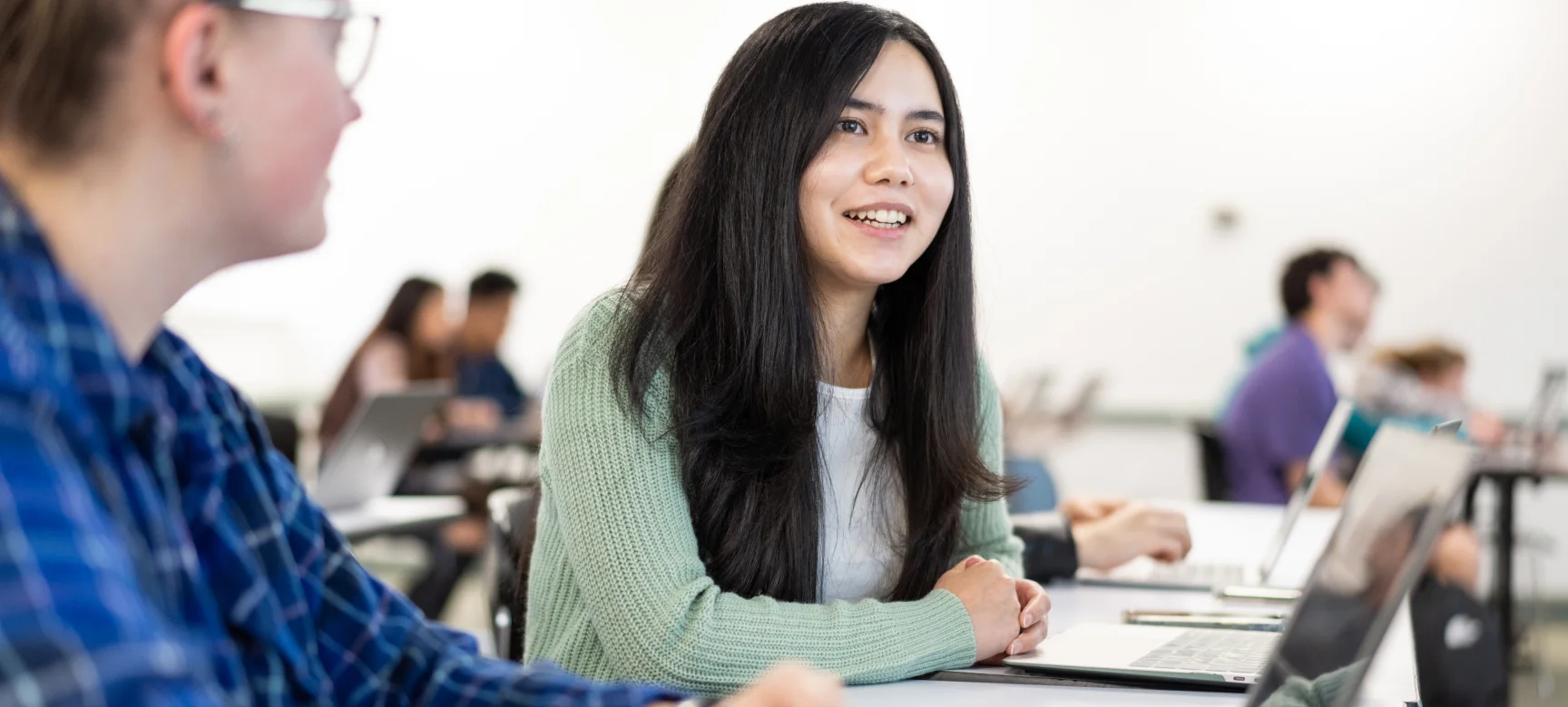 Student in a green cardigan smiling during a class discussion with laptops on desks